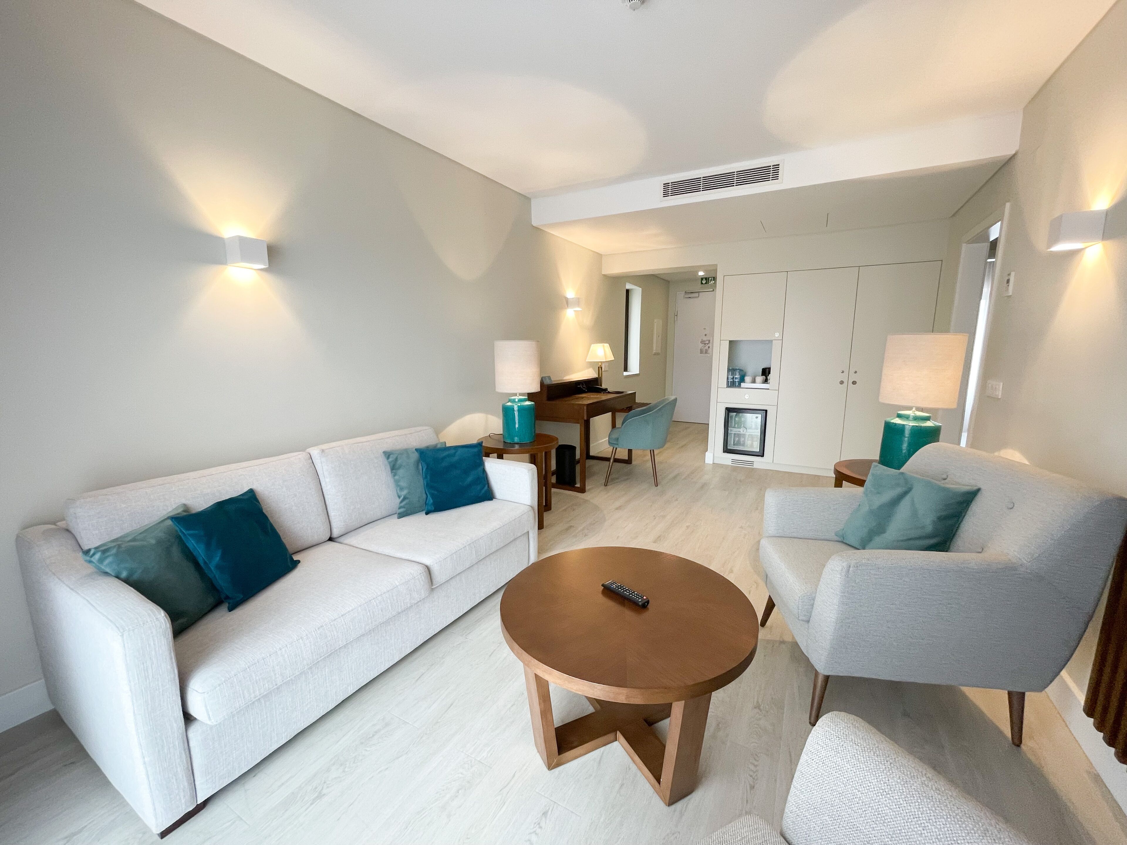 Un salon moderne avec un canapé, un fauteuil et une table basse. On y trouve également un coin repas avec une table et des chaises, ainsi qu’une petite cuisine équipée de placards et d’un réfrigérateur.