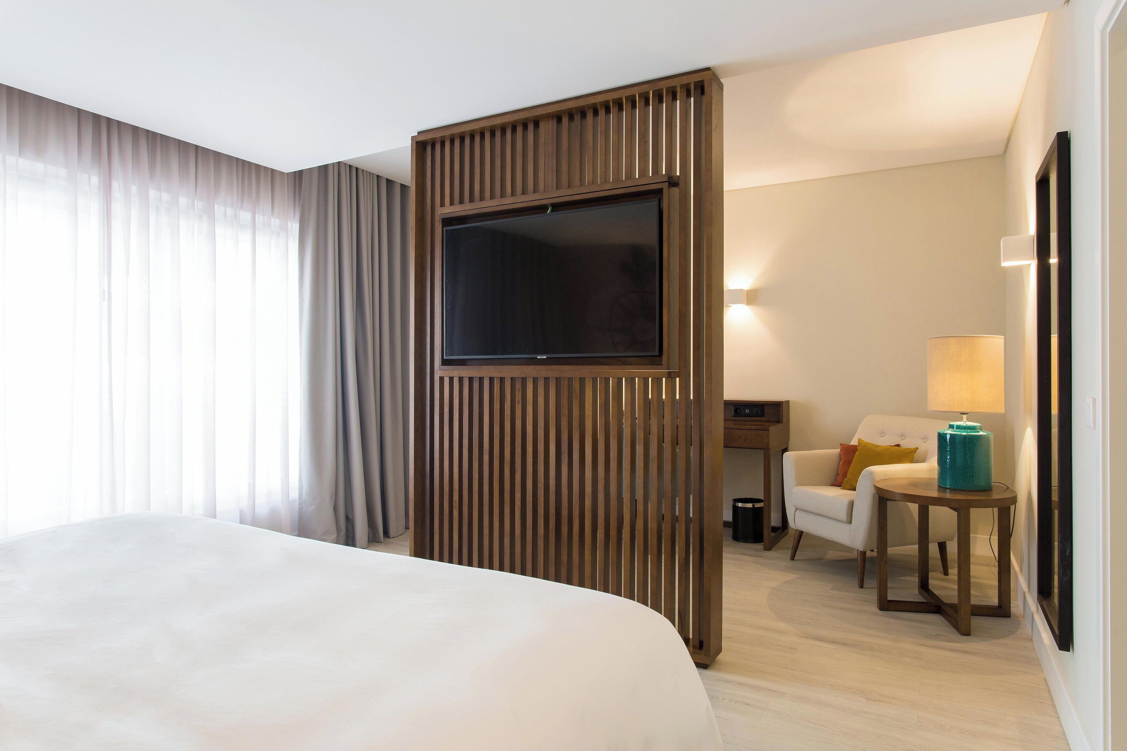 Een hotelkamer met een groot bed, een televisie bevestigd aan een houten paneel, een bureau met een lamp en een stoel.
