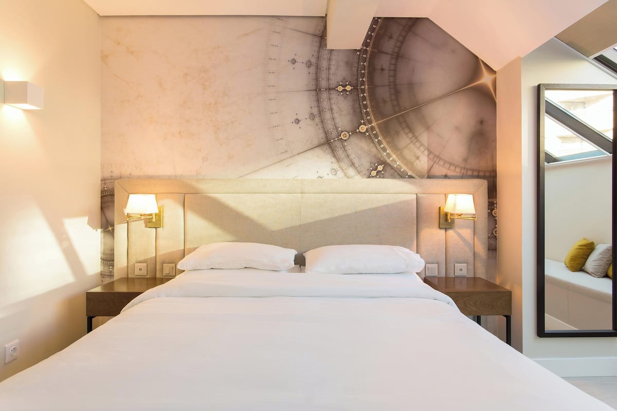 A modern hotel room with a large bed, two bedside tables with lamps, a large mirror, and a unique wall mural.