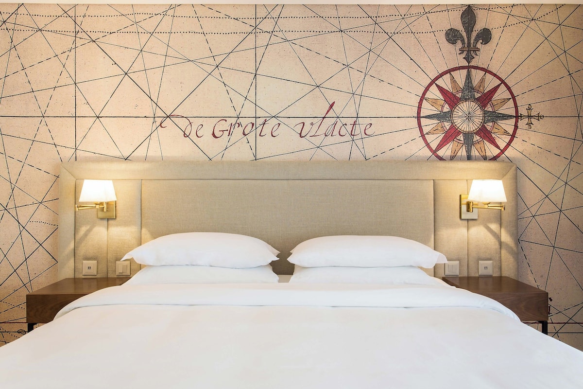 A neatly made bed with white linens, two bedside lamps, and a wall with a geometric pattern and a compass design.