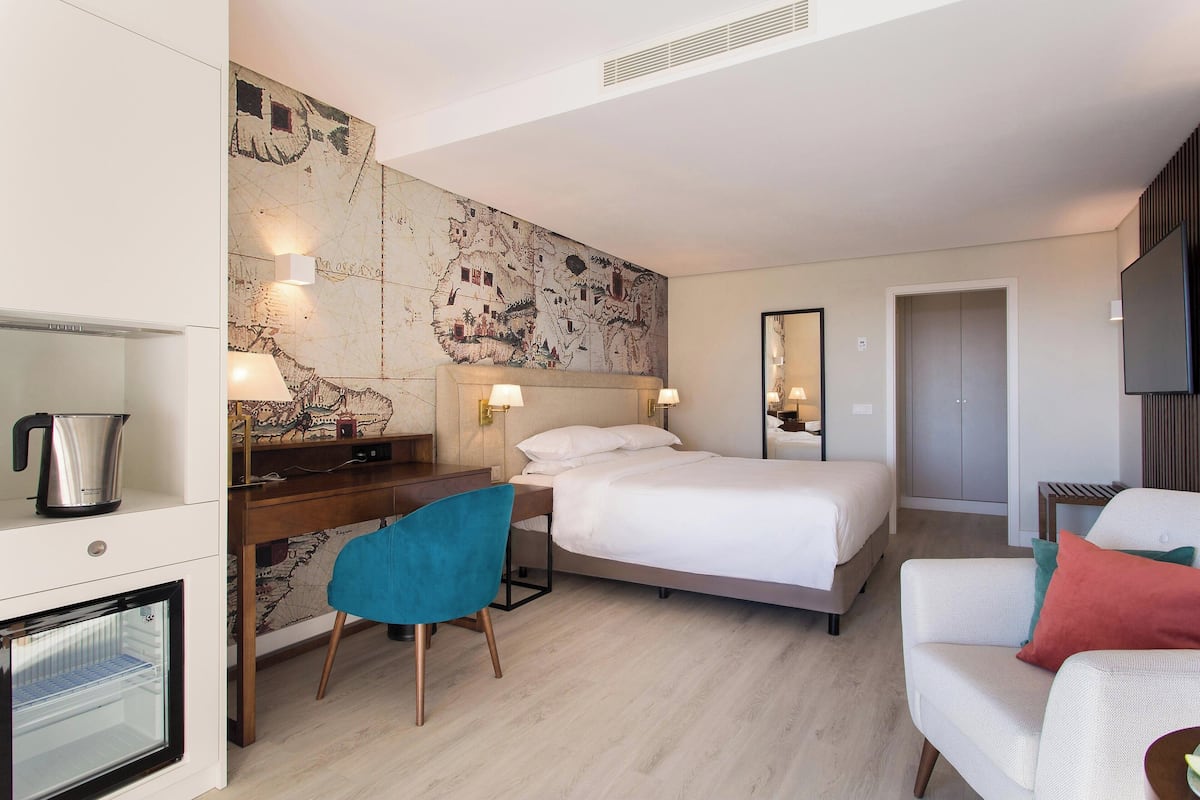 A hotel room with a bed, a desk with a chair, a minibar, and a large wall map.