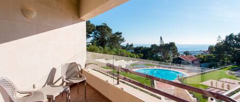 A balcony with two chairs, a view of a swimming pool, and a clear sky.
