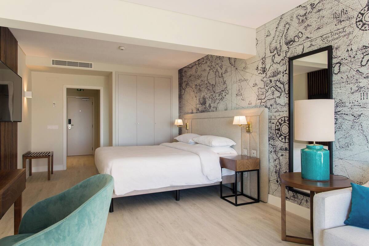 A hotel room with a bed, a desk, a chair, a mirror, and a wall with a map design.