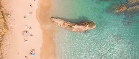 An aerial view of a beach with clear turquoise water, a rocky outcrop, and people relaxing on the sand.