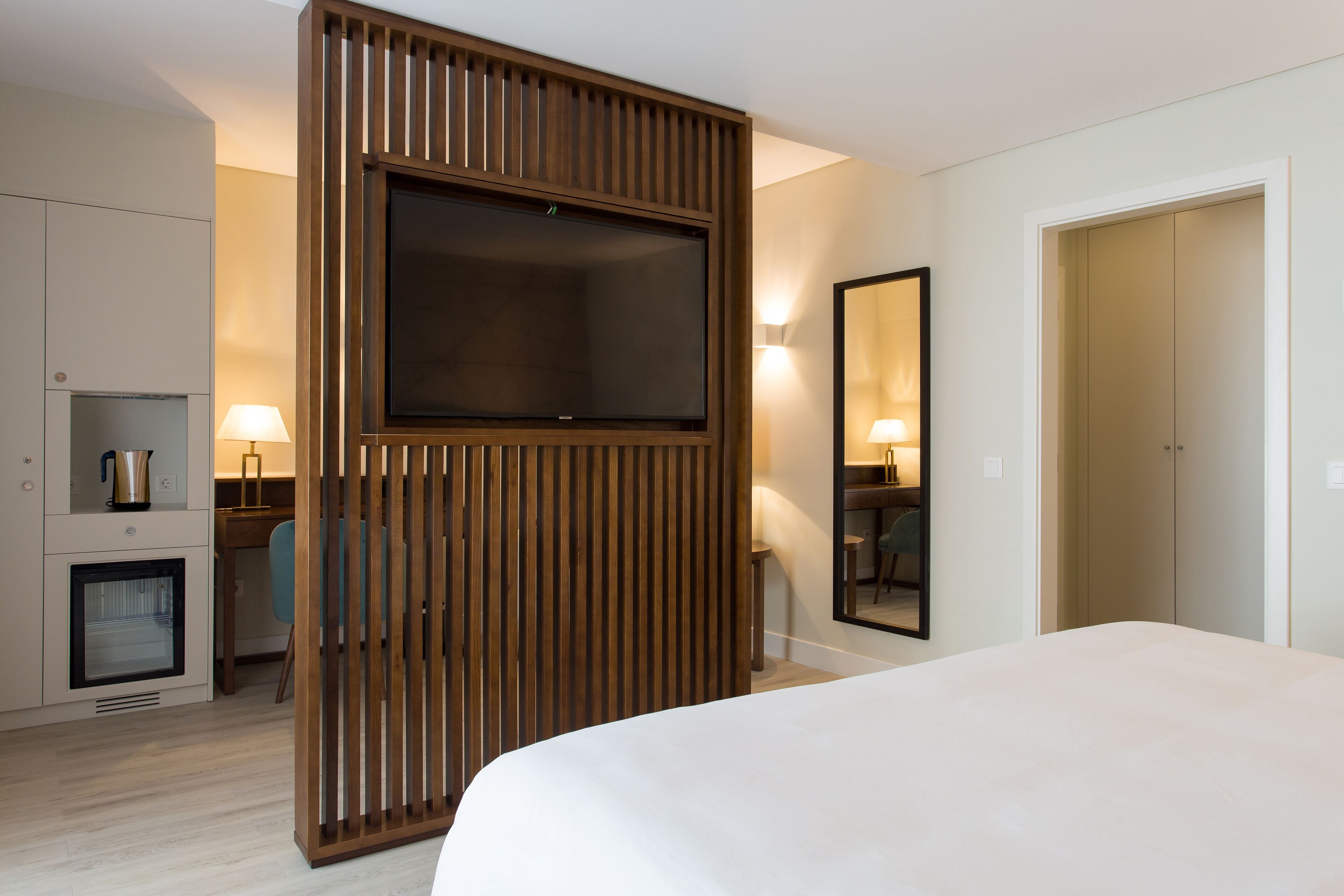 A hotel room with a large bed, a TV mounted on a wooden panel, a desk with a lamp, a chair, a minibar, and a mirror.
