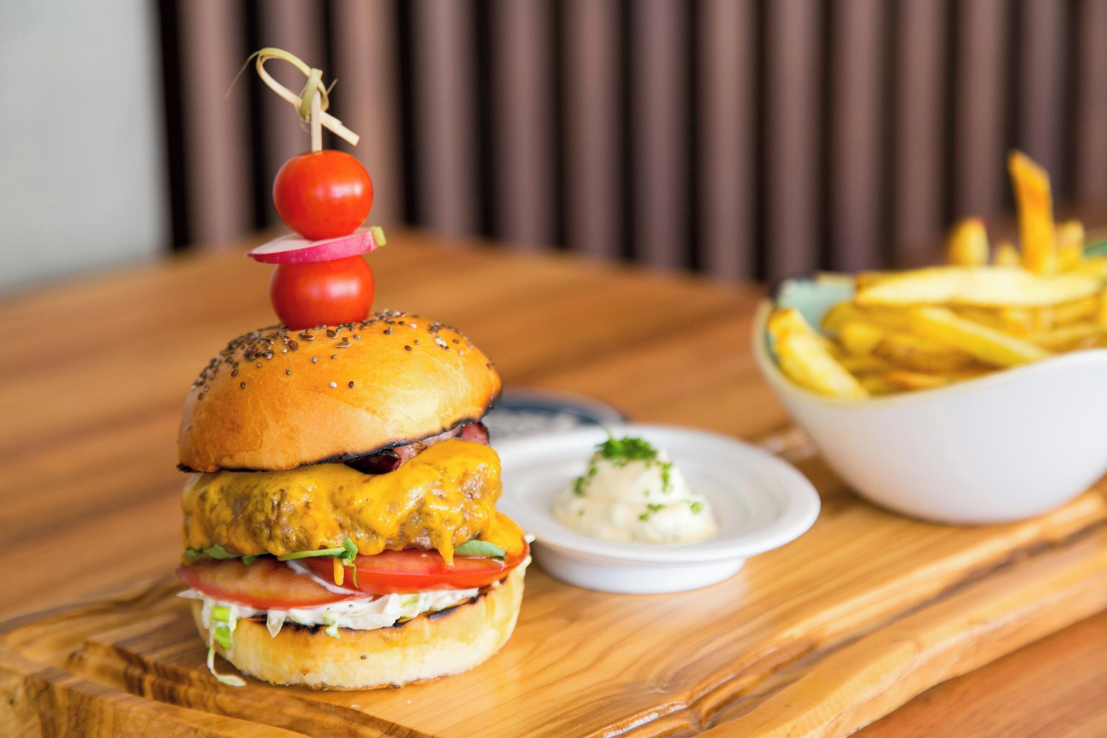 A burger with a tomato and radish on top, served with fries and a small bowl of sauce on a wooden table.
