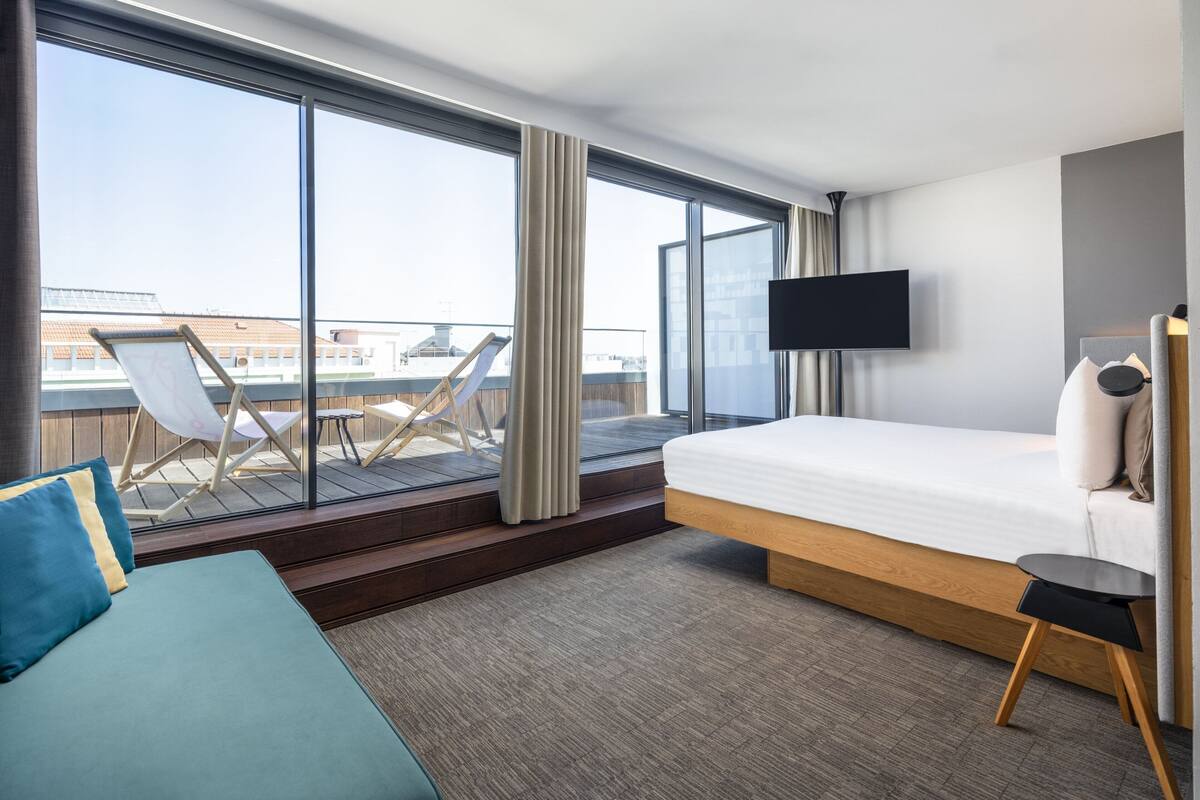 Een moderne hotelkamer met een groot raam, een bed, een televisie en een balkon met stoelen.