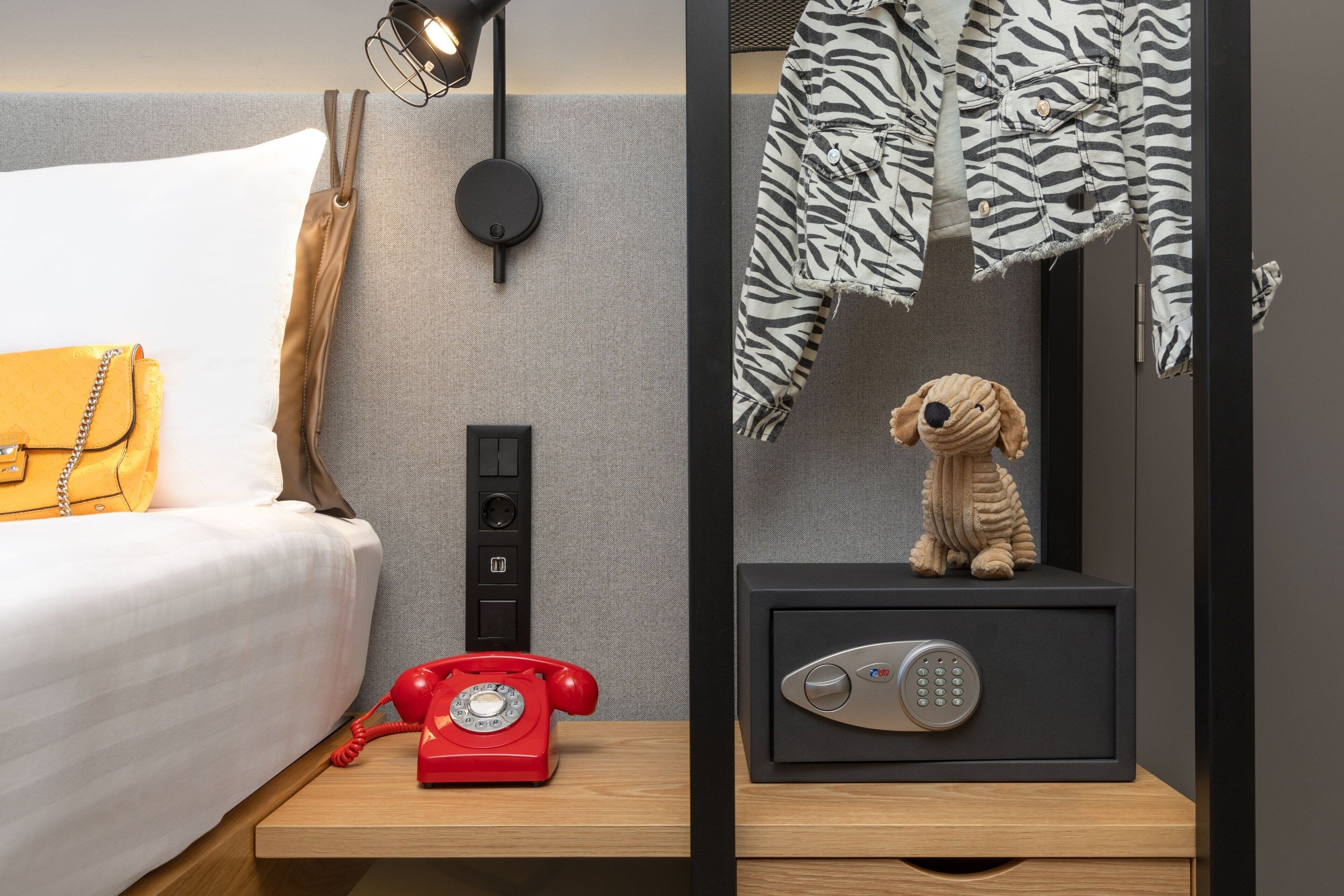 Une chambre d’hôtel avec un lit, un téléphone à cadran rouge, un coffre-fort et une veste à motifs zébrés accrochée au mur.