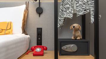 Une chambre d’hôtel avec un lit, un téléphone à cadran rouge, un coffre-fort et une veste à motifs zébrés accrochée au mur.