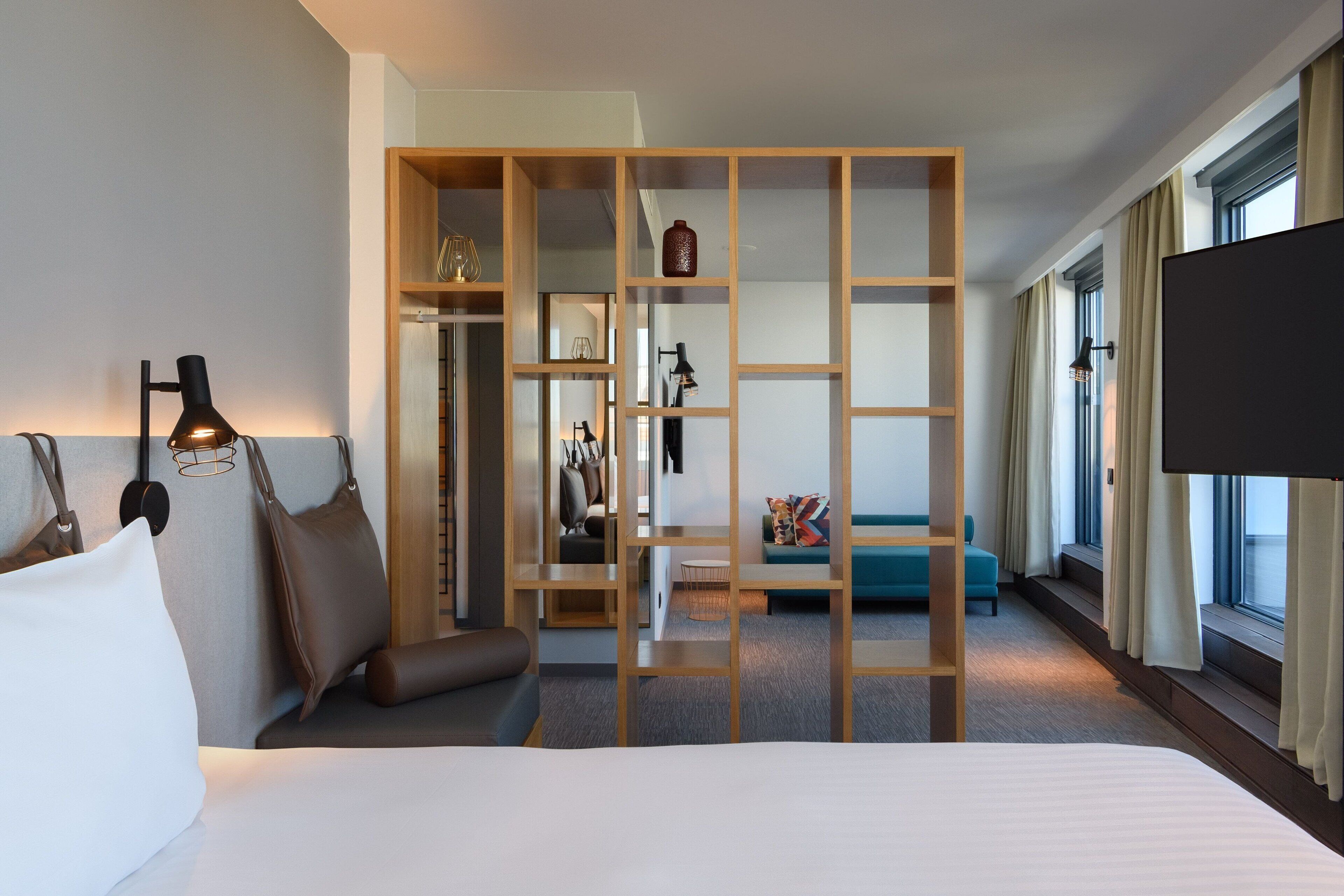 A hotel room with a bed, a wooden partition, a TV, and a view of the outside through large windows.