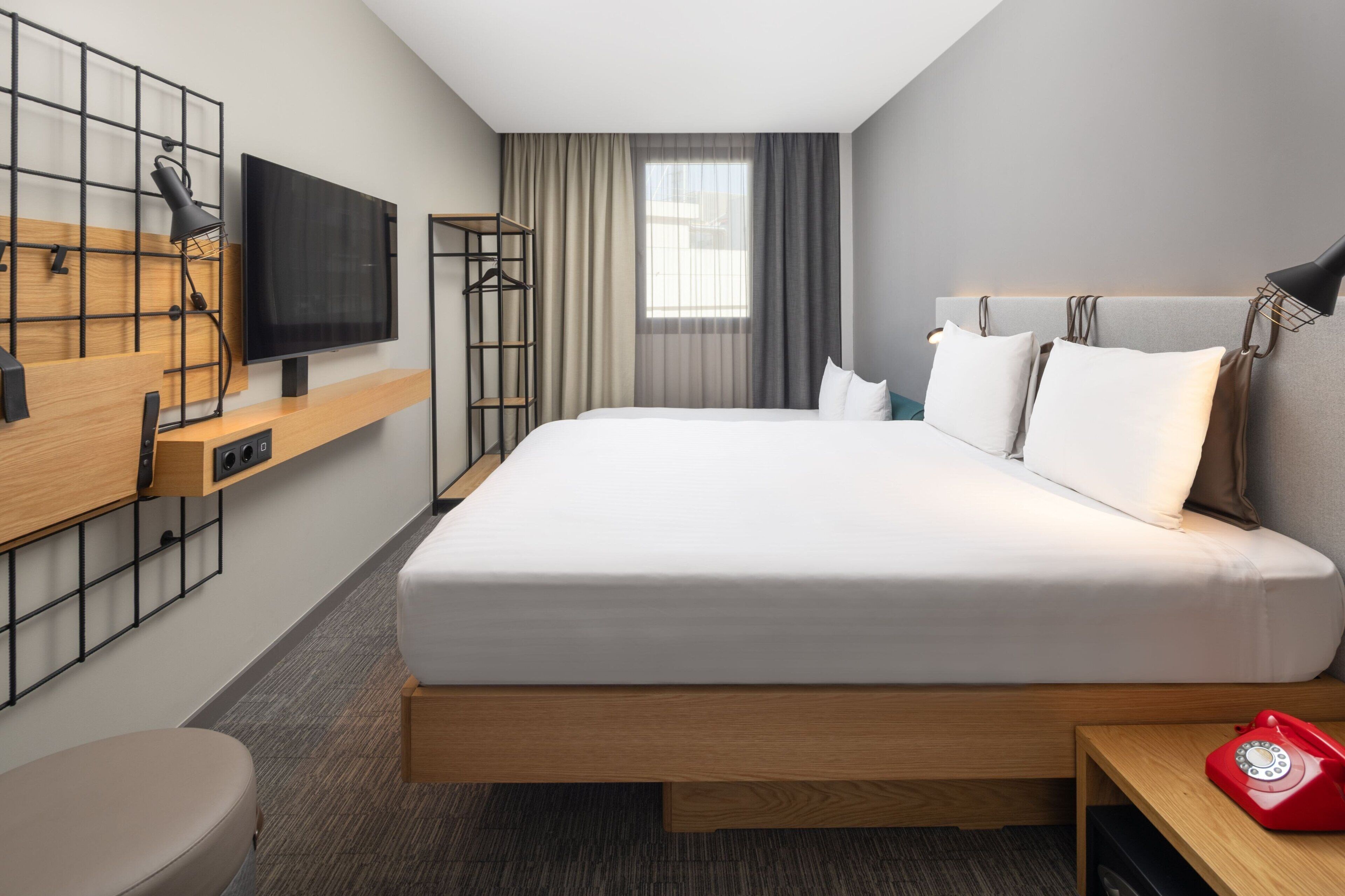 A modern hotel room with a large bed, a TV mounted on the wall, a wooden desk, a red telephone, and a window with curtains.