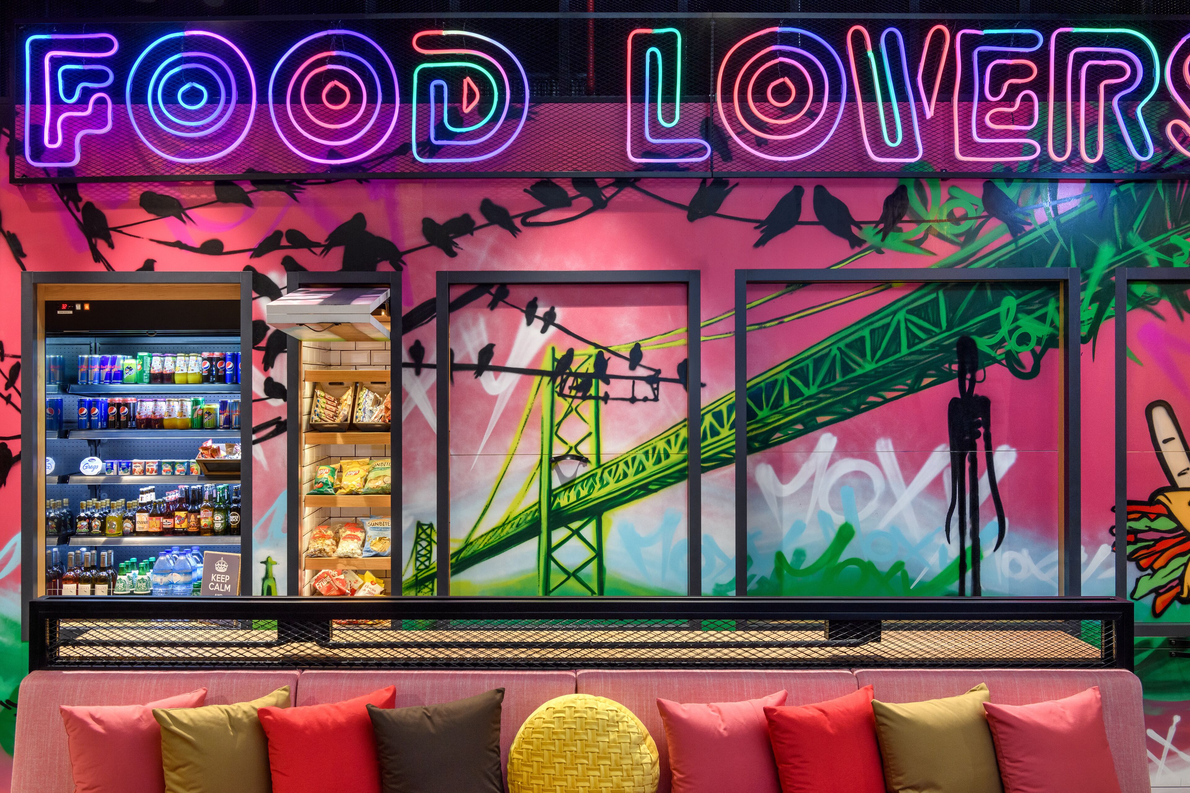 Un interior de restaurante con temática gastronómica, vibrante, con un letrero de neón, un mural de un puente y una zona de estar con cojines coloridos.