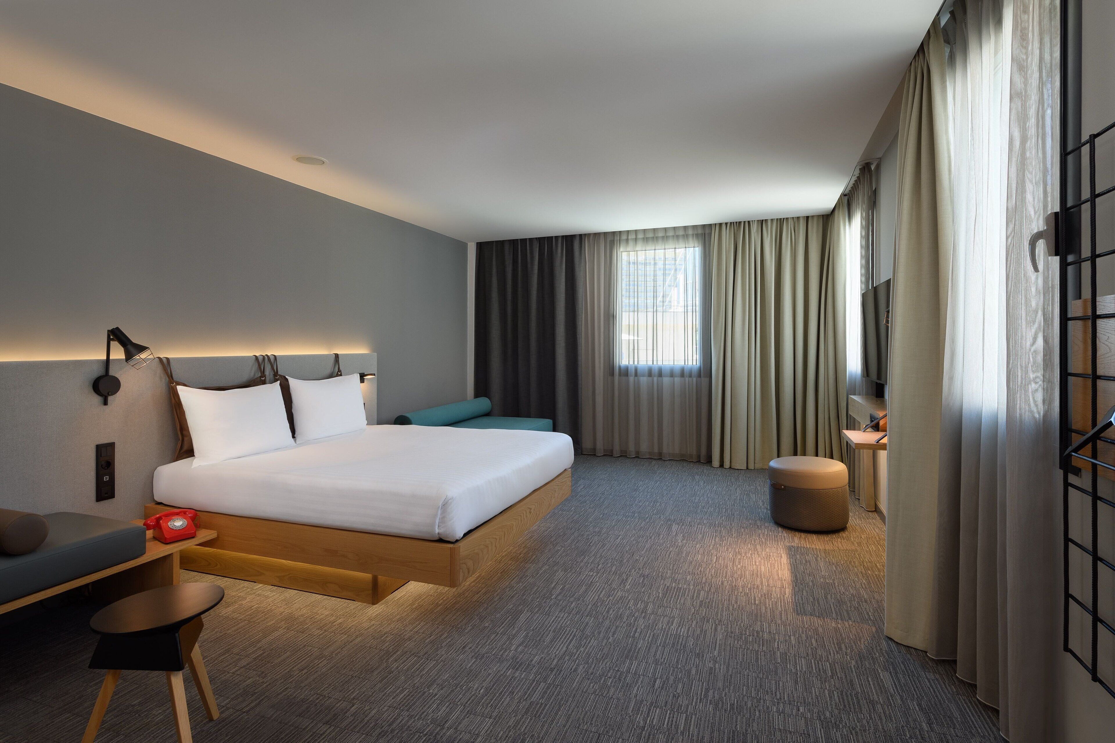 A modern hotel room with a large bed, a desk, a chair, a small table, a sofa, and a TV.