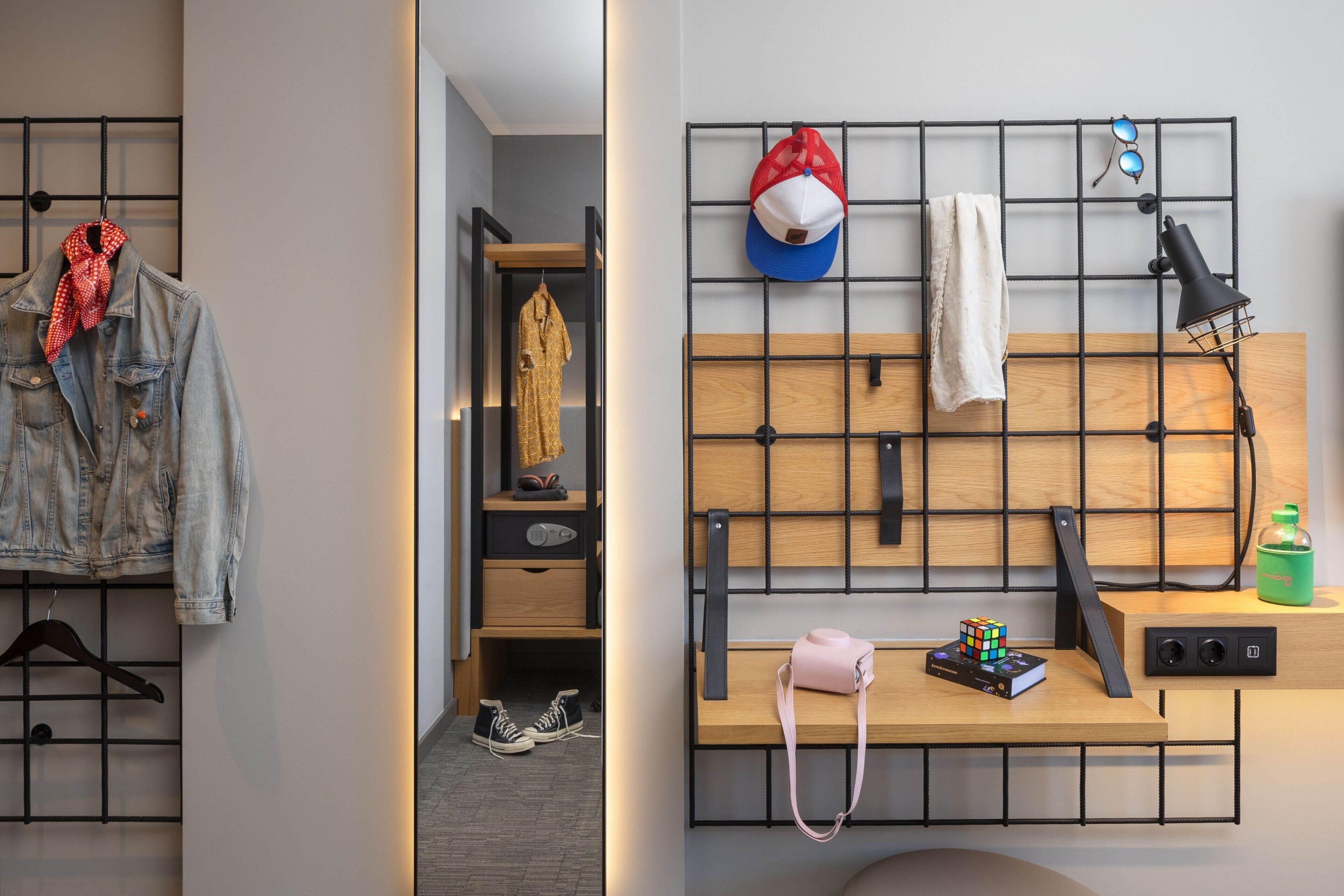 A hotel room with a wooden desk, a hanging rack with clothes, a bed with a headboard, and a wall-mounted shelf with a lamp and a Rubik's cube.