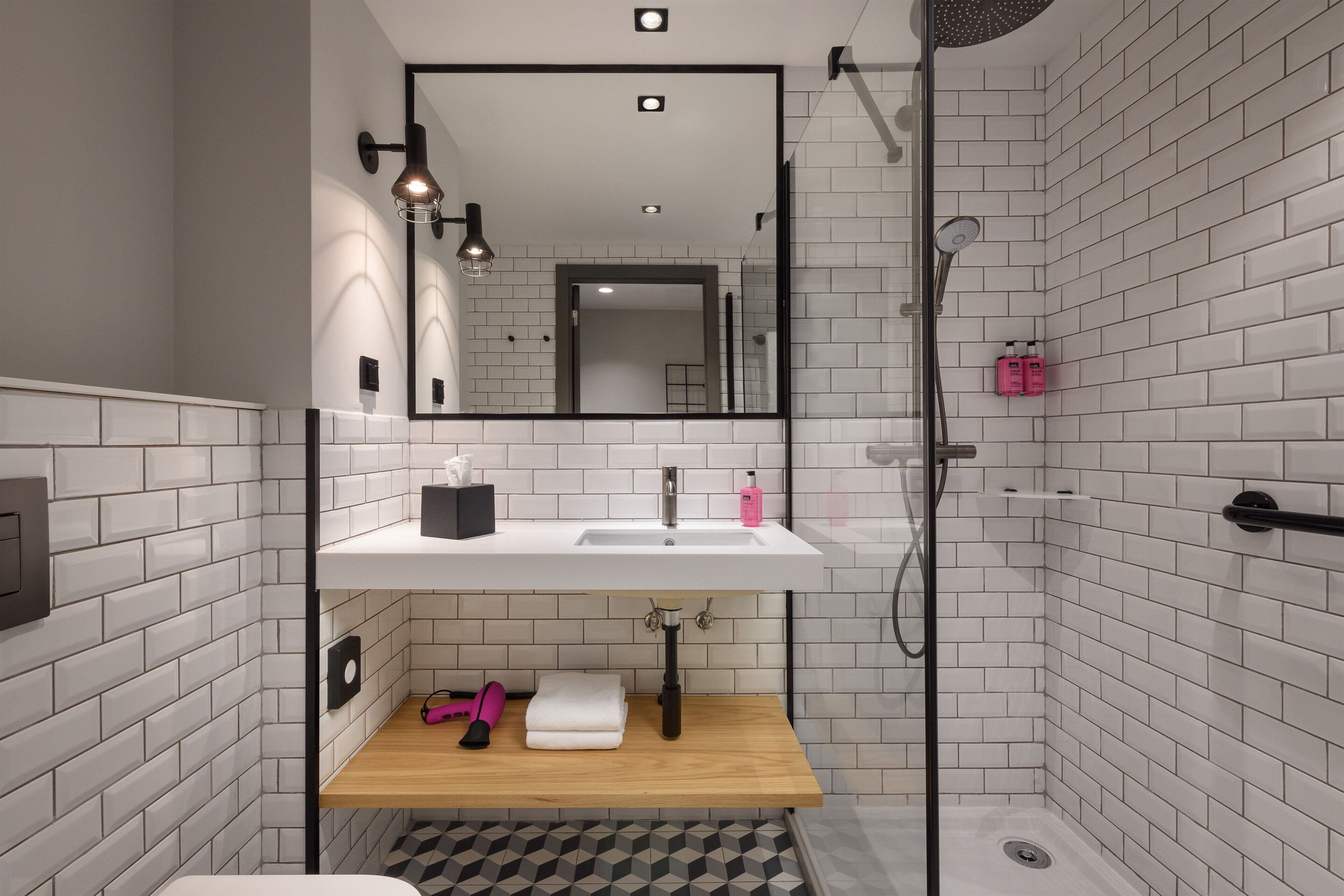 A modern bathroom with a white-tiled shower, a sink with a mirror above, and a wooden shelf with towels and toiletries.
