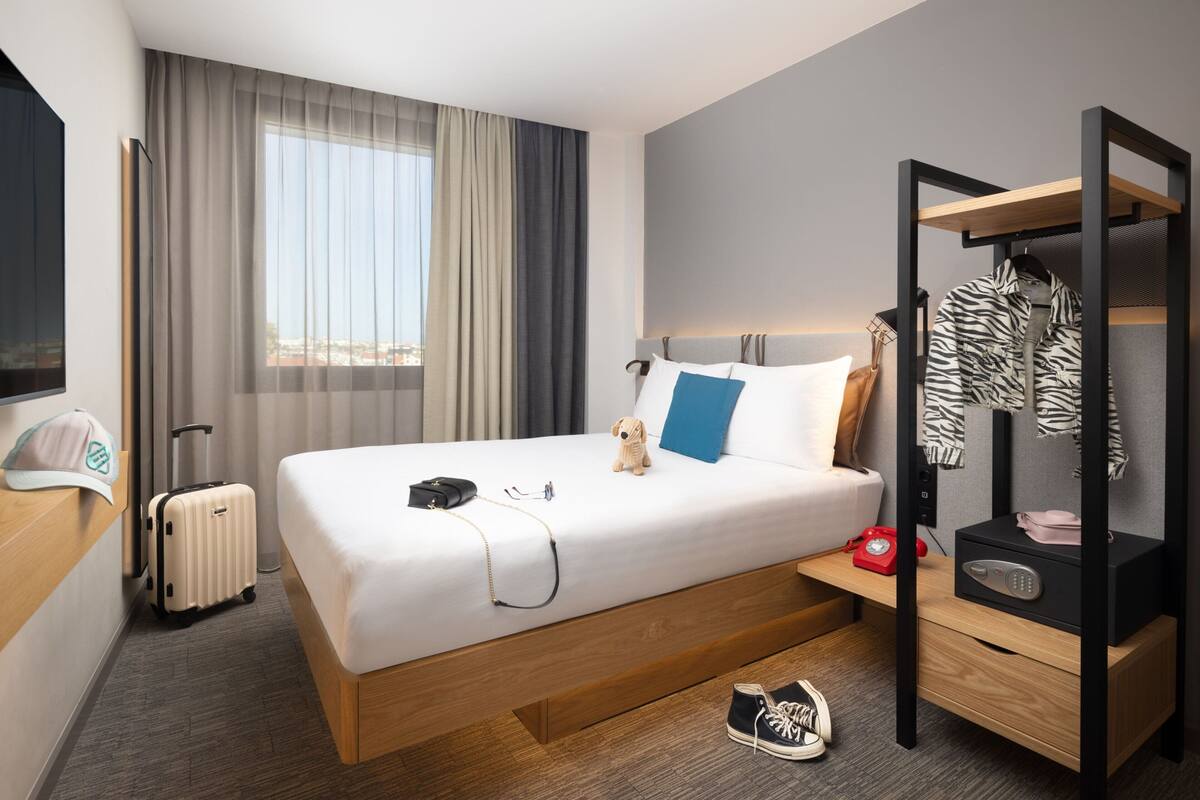 Een hotelkamer met een bed, een bureau, een koffer, een kapstok en uitzicht op de stad.