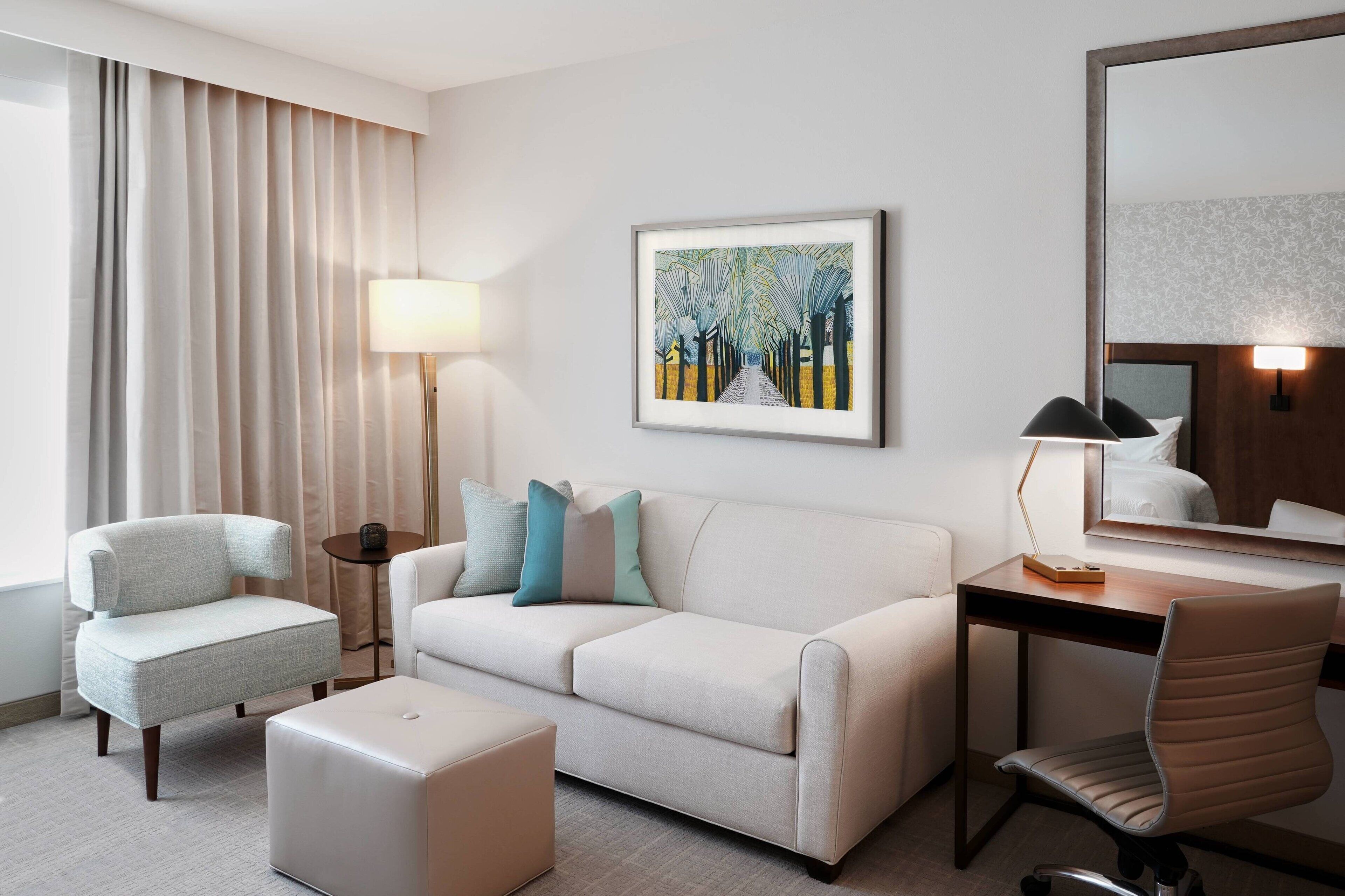 A hotel room with a sofa, armchair, desk, and chair. There is a painting on the wall and a lamp on the desk.