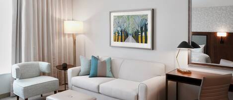 A hotel room with a sofa, armchair, desk, and chair. There is a painting on the wall and a lamp on the desk.