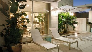 Un área de estar moderna al aire libre con dos sillones blancos y un paraguas grande, rodeada de plantas en macetas y una puerta de vidrio que da a un espacio interior.