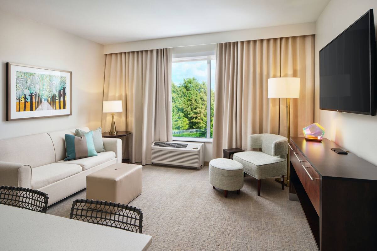 A modern hotel room with a sofa, ottoman, desk, and a large window with curtains.