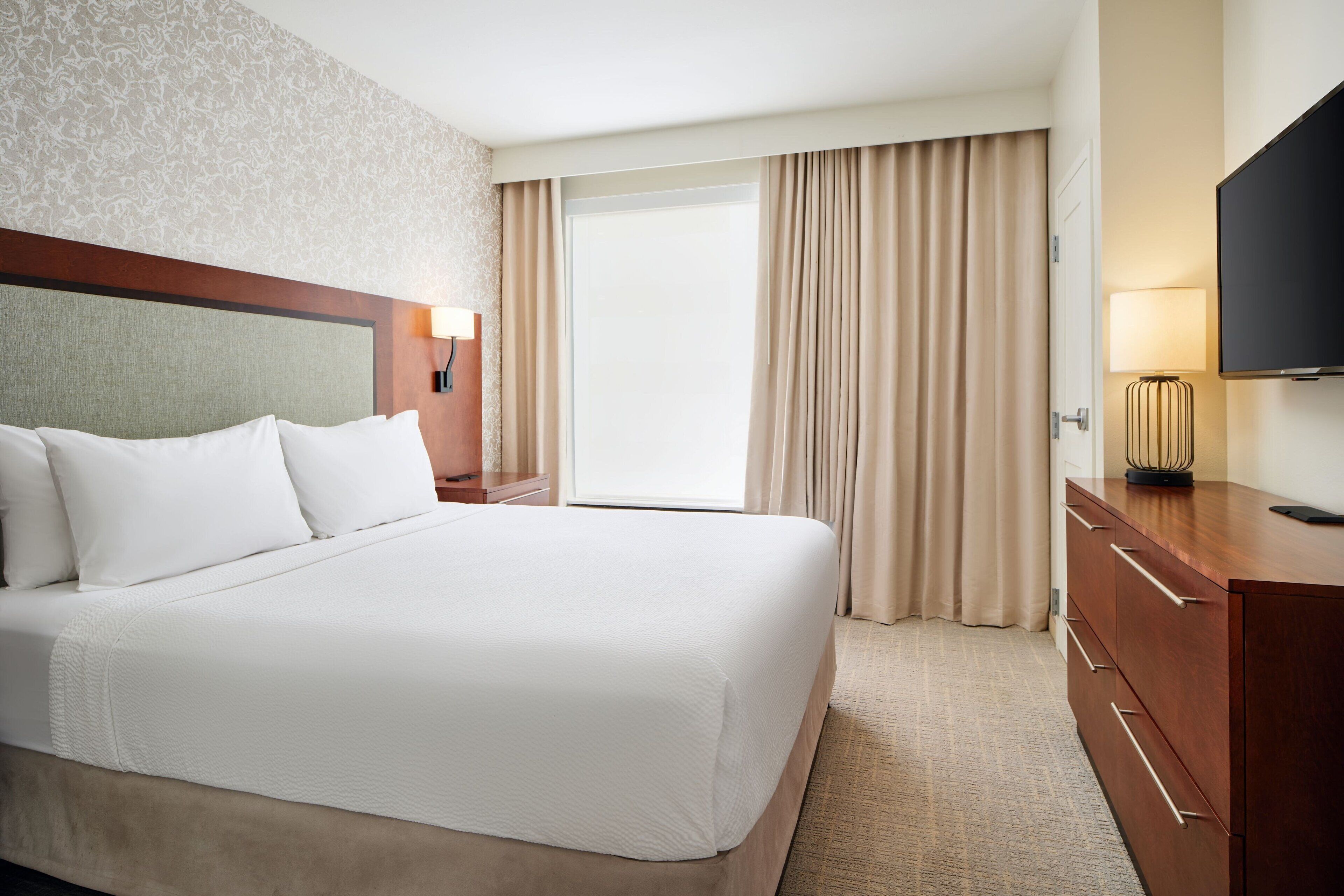 Habitación de hotel con una cama grande, cabecero de madera, televisor de pantalla plana, escritorio con cajones y ventana con cortinas.