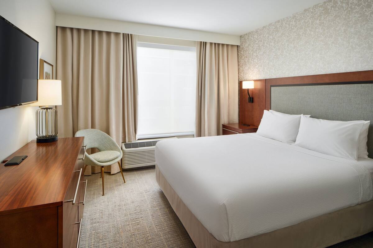A hotel room with a large bed, a desk with a chair, a TV, and a window with curtains.