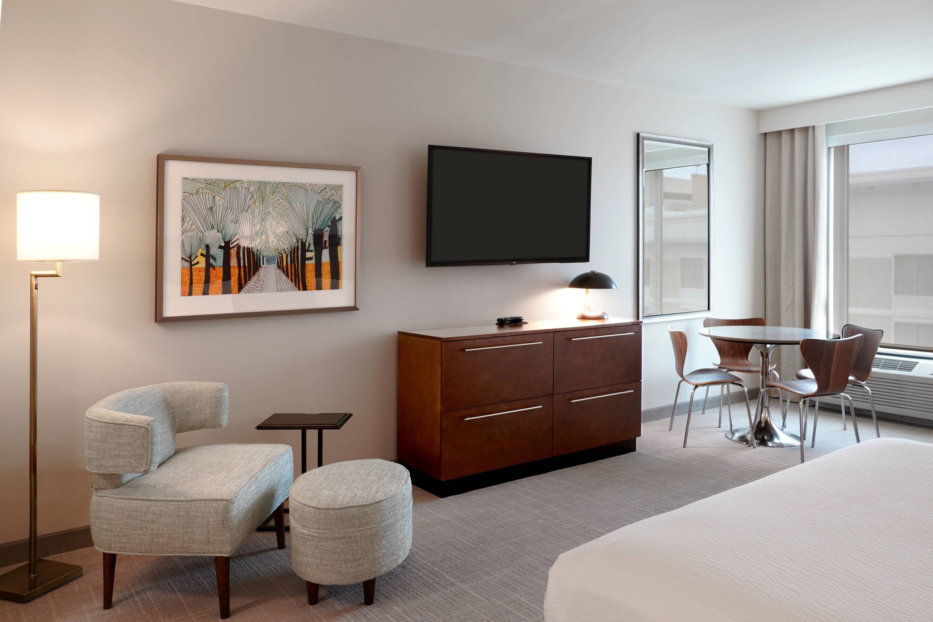 A hotel room with a bed, a desk with a chair, a television, a framed picture, and a floor lamp.