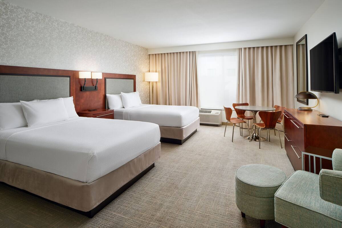 A hotel room with two beds, a desk, a chair, a TV, and a window with curtains.