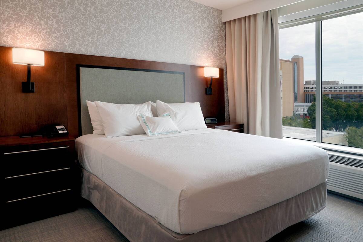 A hotel room with a large bed, two bedside lamps, a nightstand, and a view of the city through large windows.