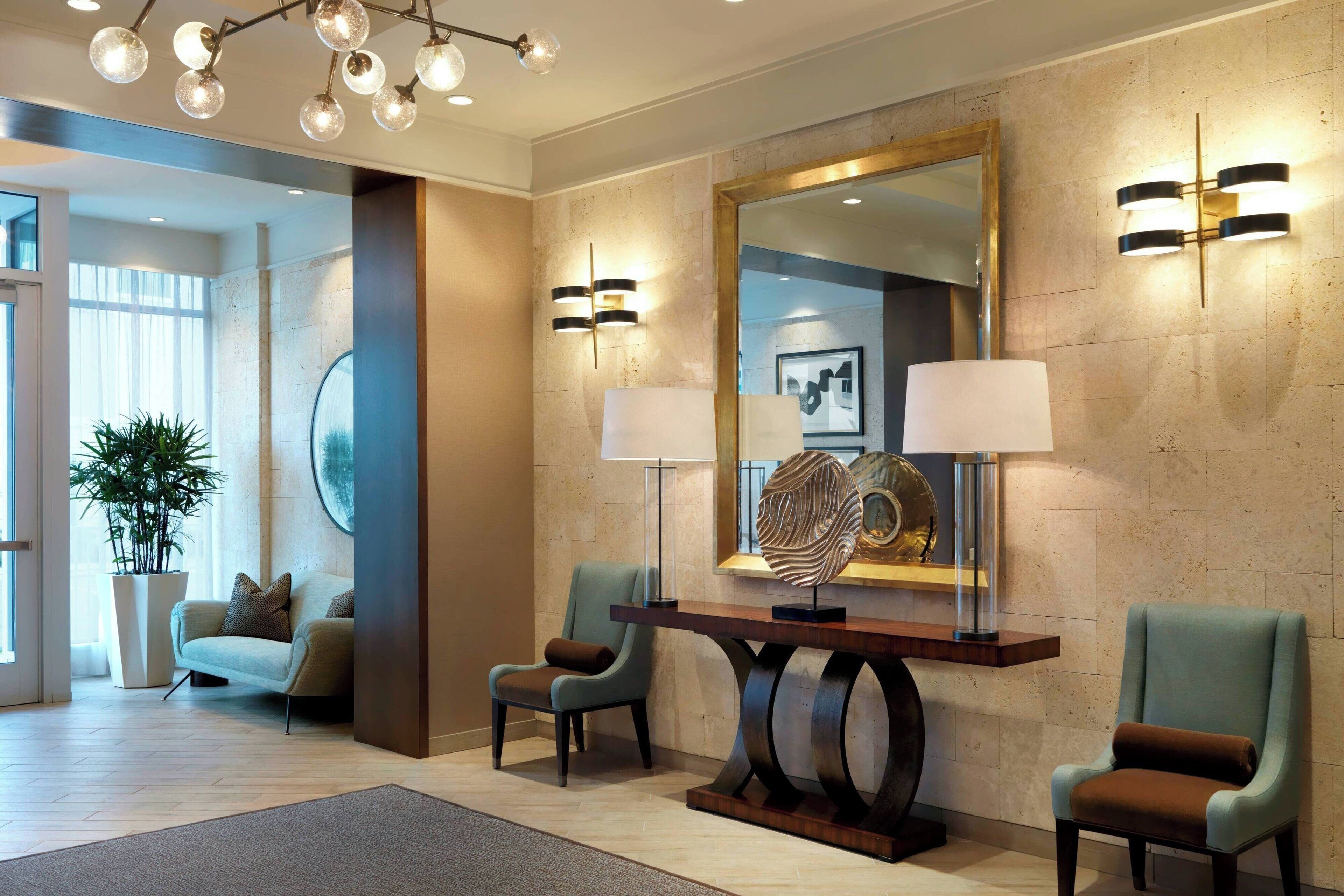A modern lobby with a large mirror, two floor lamps, a wooden console table, and two armchairs.