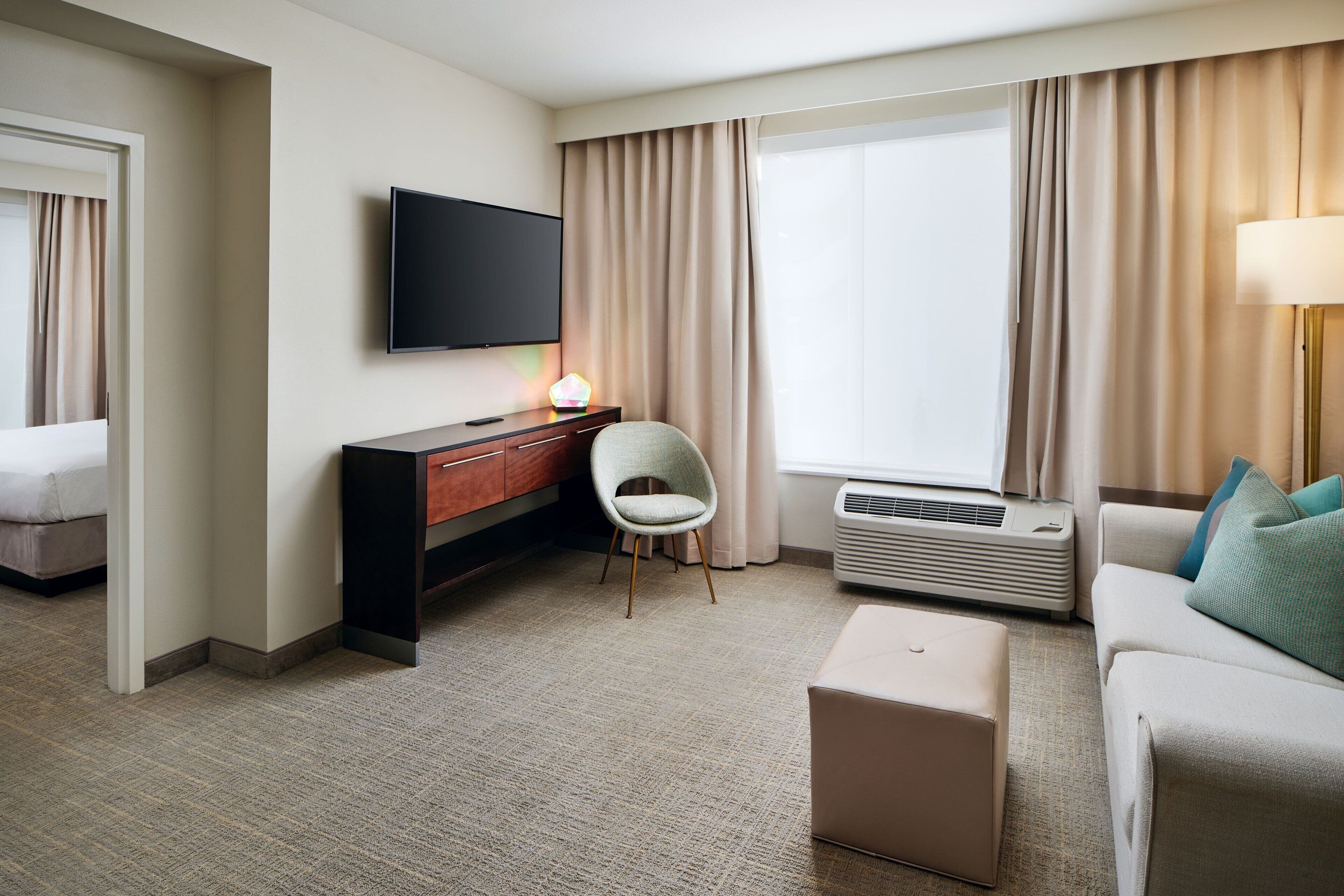 A hotel room with a flat-screen TV, a desk with a chair, a sofa, a small ottoman, and a large window with curtains.