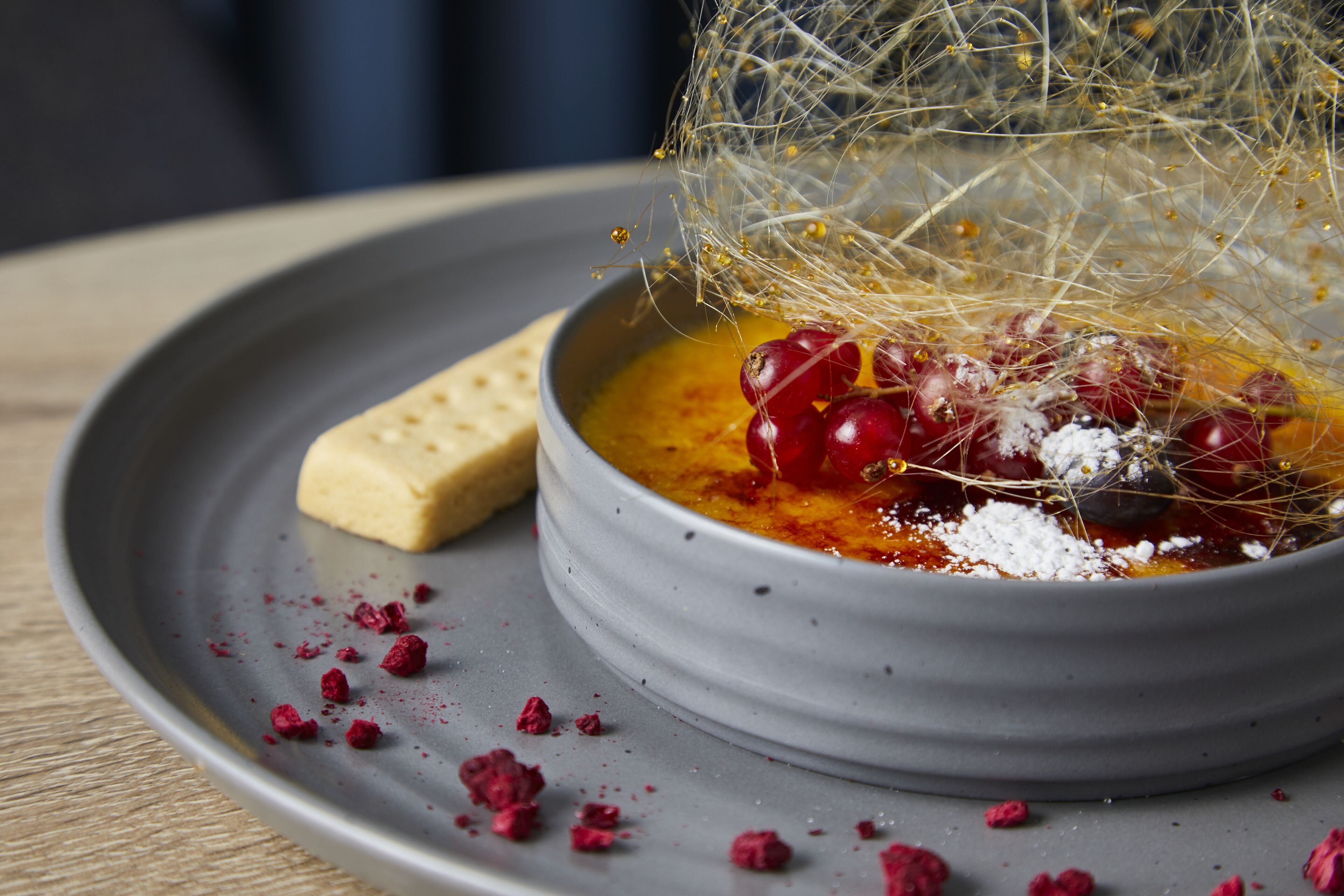 A bowl of soup with a decorative net, garnished with red berries and a crumbled topping, on a gray plate.