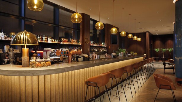 A bar area with a long counter, multiple stools, and hanging pendant lights. - Four Points by Sheraton Prishtina City (Preoce)
