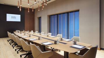 A conference room with a long table, chairs, a projector screen, and large windows with blinds.