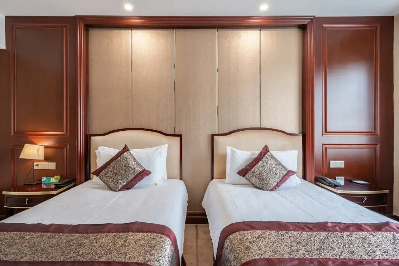 Una habitación de hotel con dos camas, cada una con colcha y almohadas estampadas, una mesita de noche con lámpara y un panel de pared de madera.