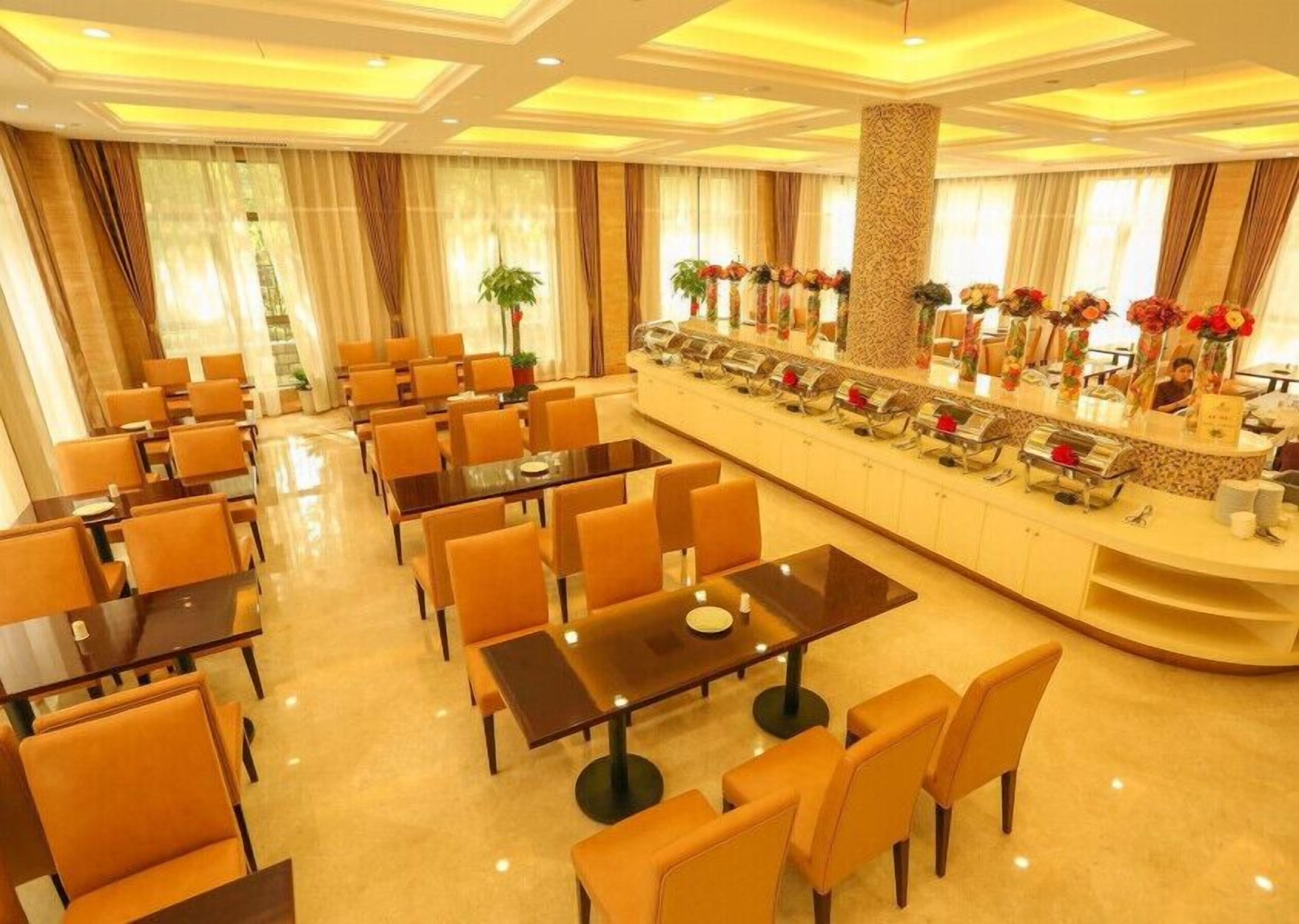 Un'area ristorazione d'albergo con diversi tavoli e sedie, un banco buffet con esposizione di cibi e un ampio pilastro al centro.