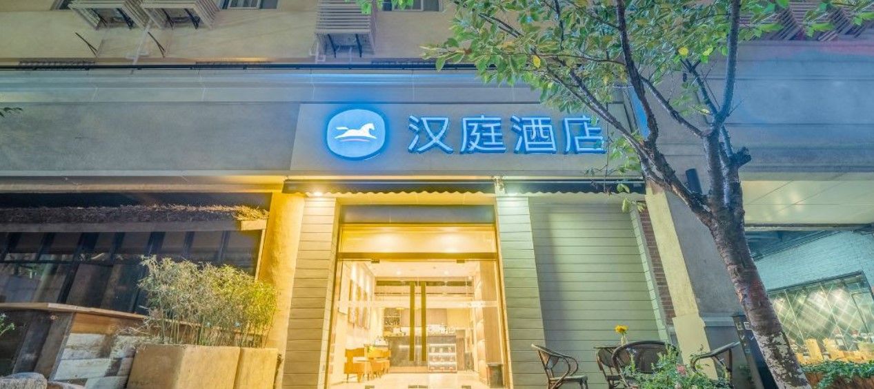 Entrada de hotel com uma placa em caracteres chineses, uma bicicleta estacionada e uma árvore em primeiro plano.