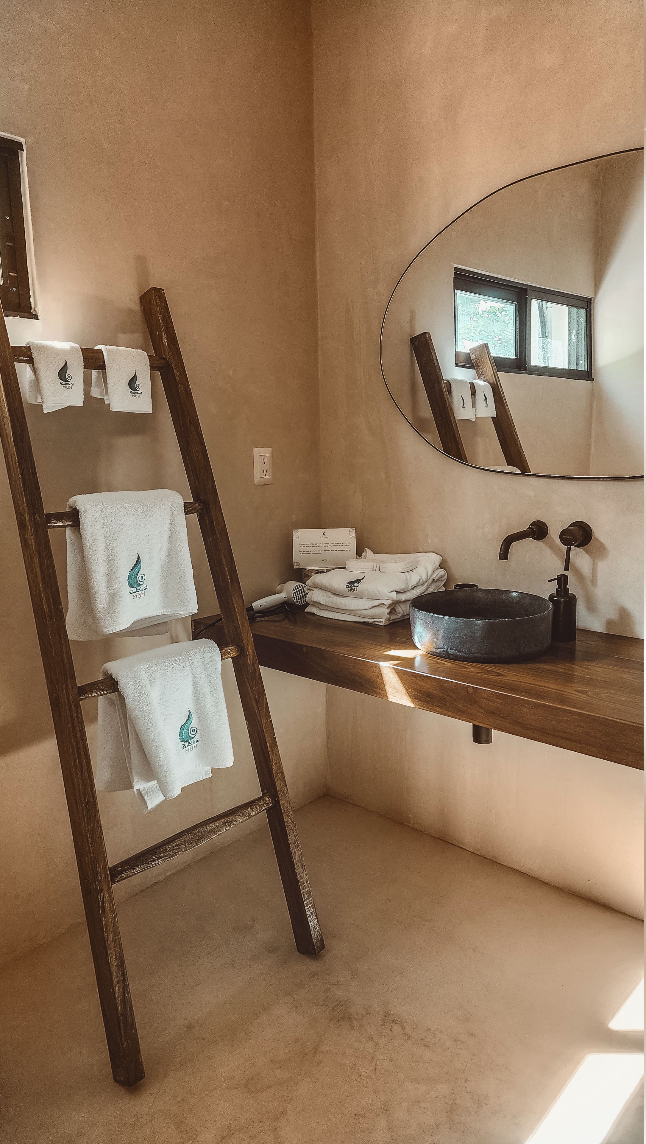A bathroom with a wooden ladder, a sink with a round basin, a mirror, and towels with a water droplet logo.