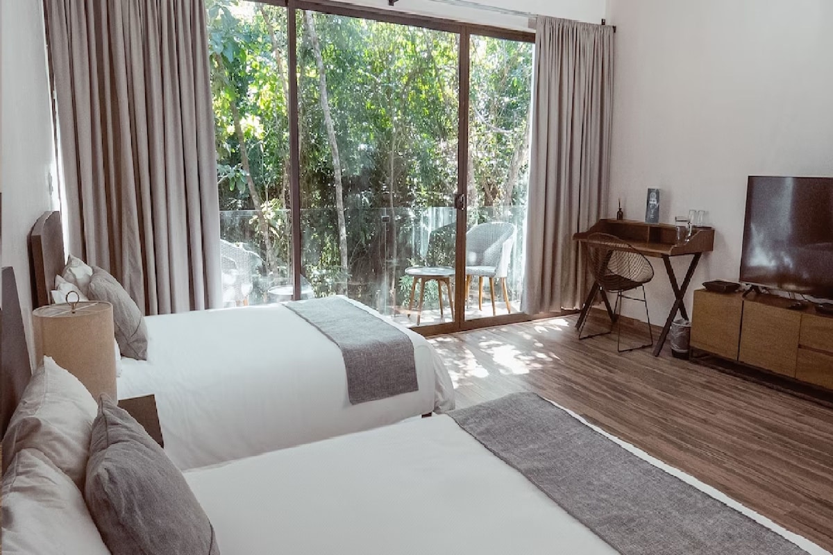 A bedroom with a large bed, a desk, a TV, and a view of greenery through a sliding glass door.