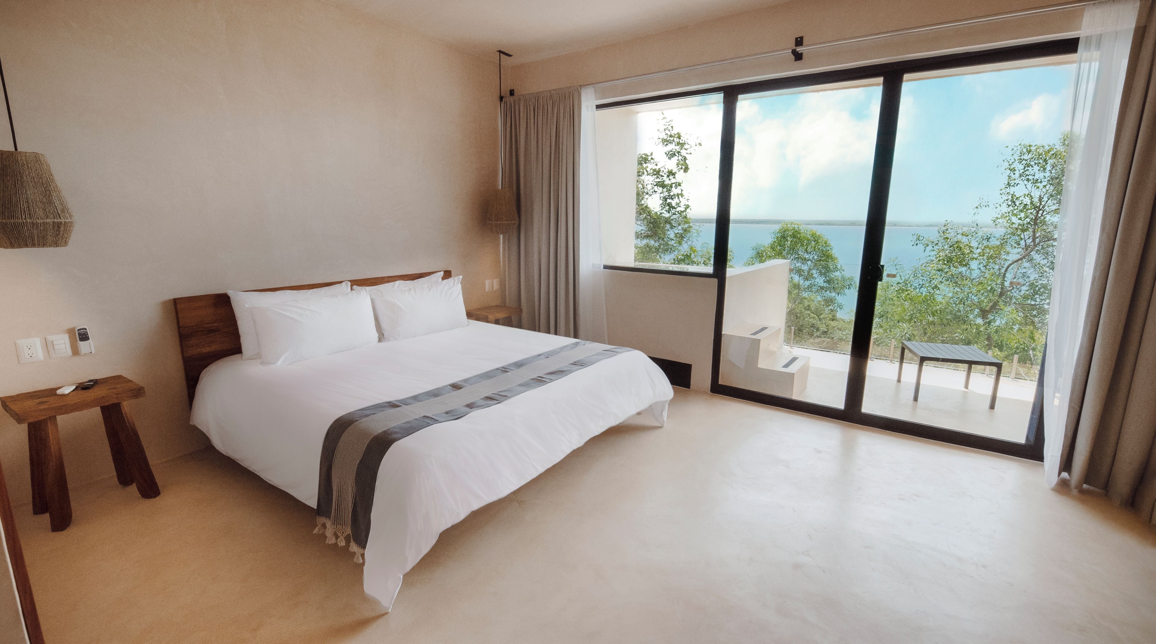 Una habitación de hotel moderna con una cama grande, una mesita de noche de madera, una lámpara y vistas al mar a través de una puerta corrediza de vidrio.