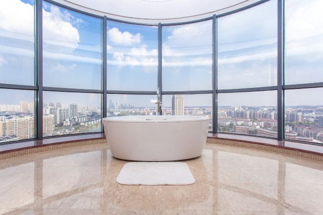 A modern bathroom with a large bathtub, a white rug, and panoramic city views through large windows.
