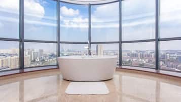 A modern bathroom with a large bathtub, a white rug, and panoramic city views through large windows.
