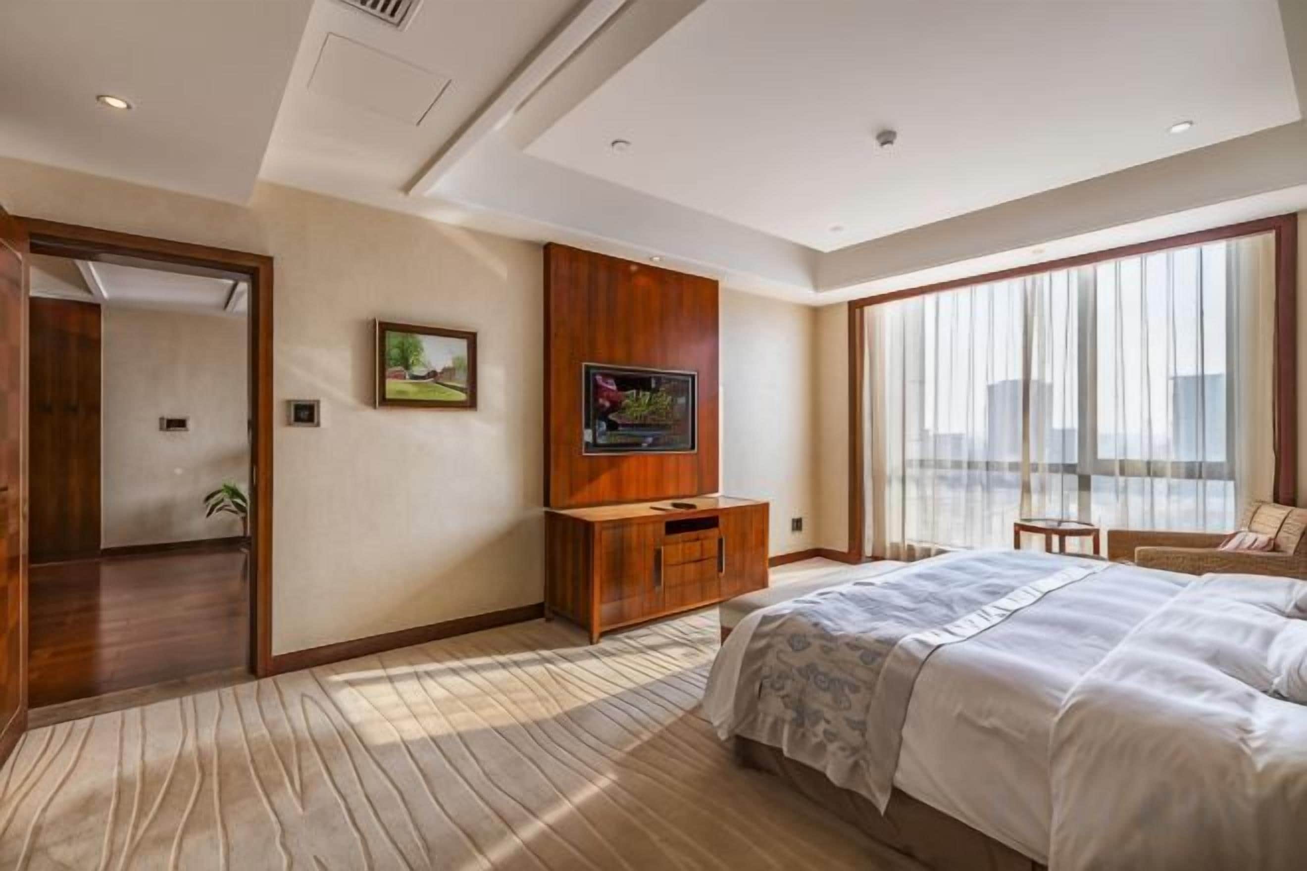 Habitación de hotel con una cama grande, televisor, un aparador de madera y vistas de la ciudad a través de amplios ventanales.