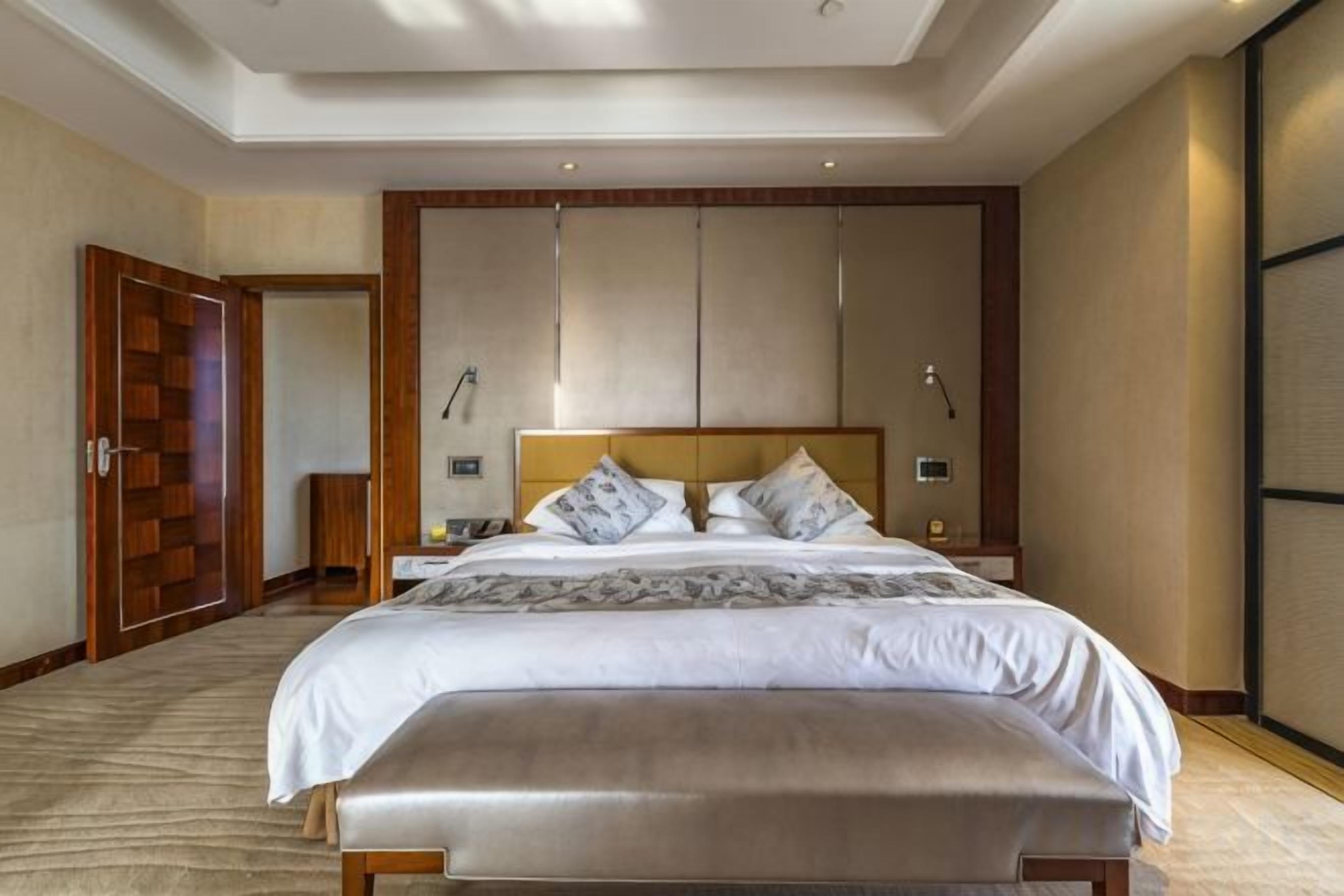 A hotel room with a large bed, a bedside table, a bench, and a wardrobe.
