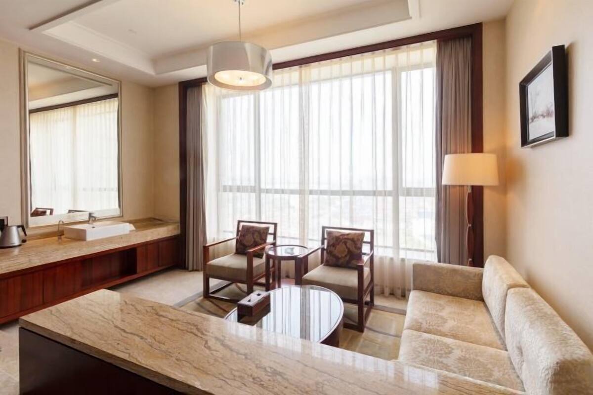 A hotel room with a marble countertop, two chairs, a sofa, a round table, a large window with curtains, a mirror, and a wall-mounted lamp.