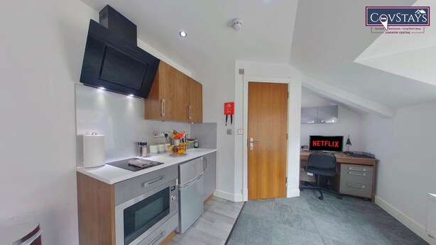 A modern kitchen with a built-in oven, microwave, and a flat-screen TV. There's a small office area with a desk and a computer. - Victoria House - Deluxe Studio by CovStays (Coventry)