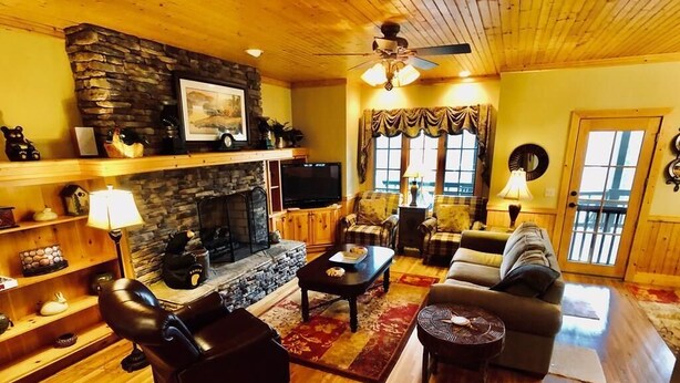 A cozy living room with a stone fireplace, wooden floors, a ceiling fan, and a large window with curtains.