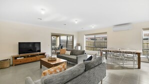 A spacious living room with a flat-screen TV, grey sofas, a wooden coffee table, and a dining area with a table and chairs. - Ocean View Crescent 59 Torquay Pet Friendly (TORQUAY)