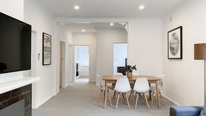 A modern living room with a flat-screen TV, a dining area with a wooden table and white chairs, and a hallway leading to another room. - Immaculately prepared property is only 40 metres from the foreshore (TORQUAY)