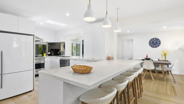 A modern kitchen with white cabinetry, a large island with bar stools, pendant lighting, and a clock on the wall. - Puebla Casa 17  Walk to Beach & Shops (TORQUAY)